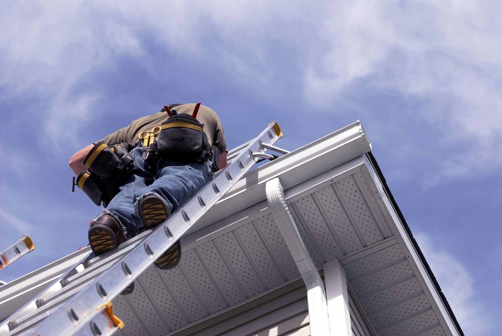 Roofer in a tool belt climbing an aluminum ladder to the eave of a house with white gutters and a downspout